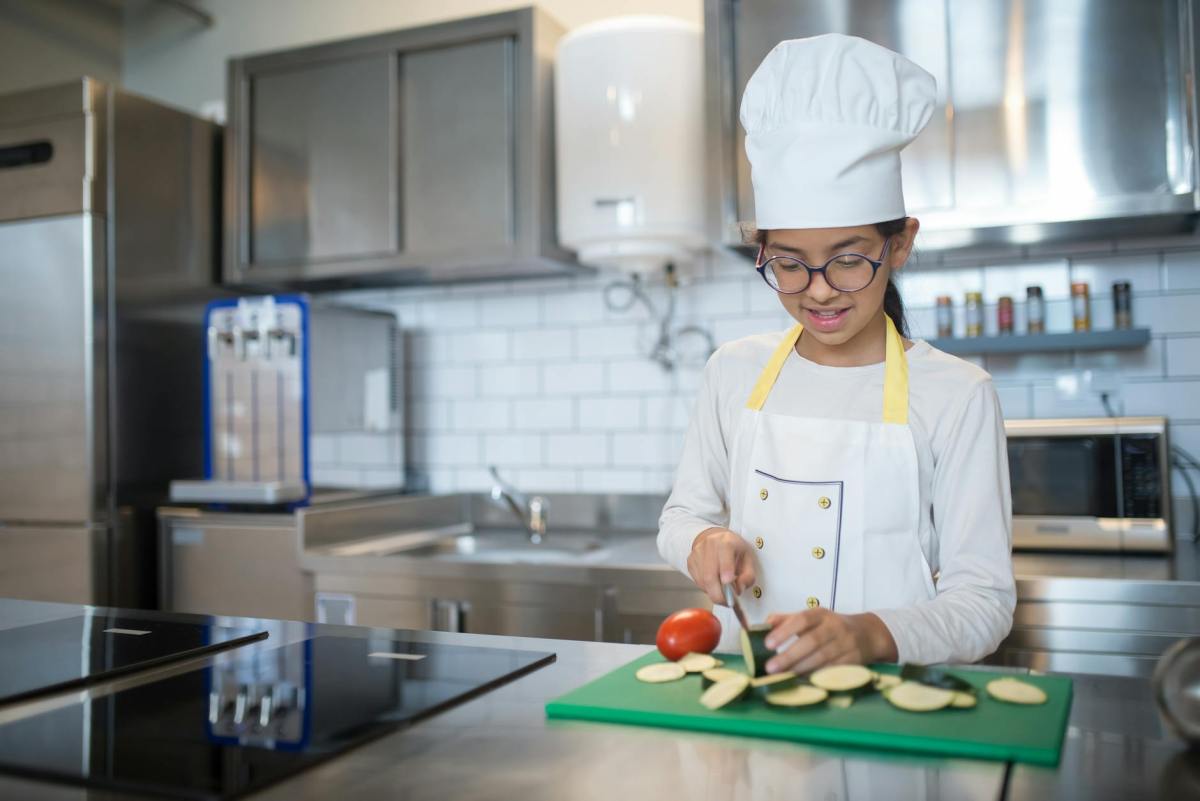 a girl in white apron and chef s hat slicing a cucumber on a chopping board