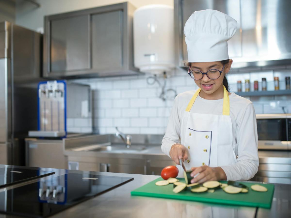 a girl in white apron and chef s hat slicing a cucumber on a chopping board
